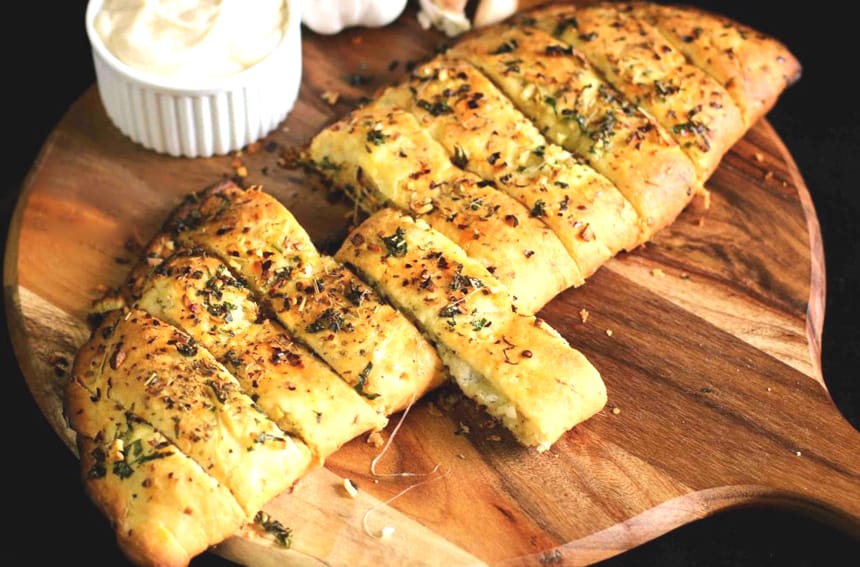 A photo of freshly baked garlic bread, sliced and spread with melted cheese, garnished with chopped parsley, and served on a white plate.