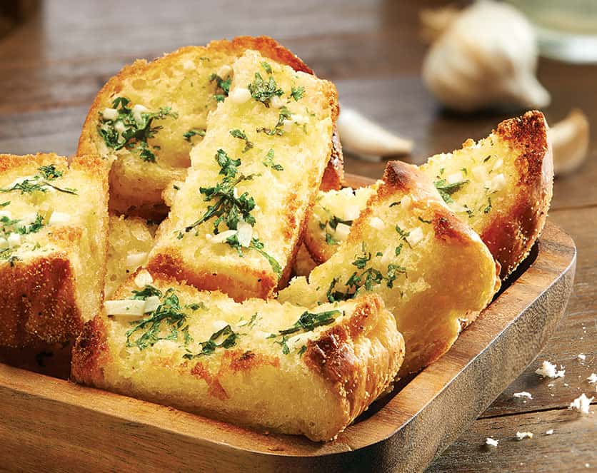 A photo of a plate of garlic bread, with four slices of bread brushed with garlic butter and sprinkled with herbs, served with a small bowl of marinara sauce.