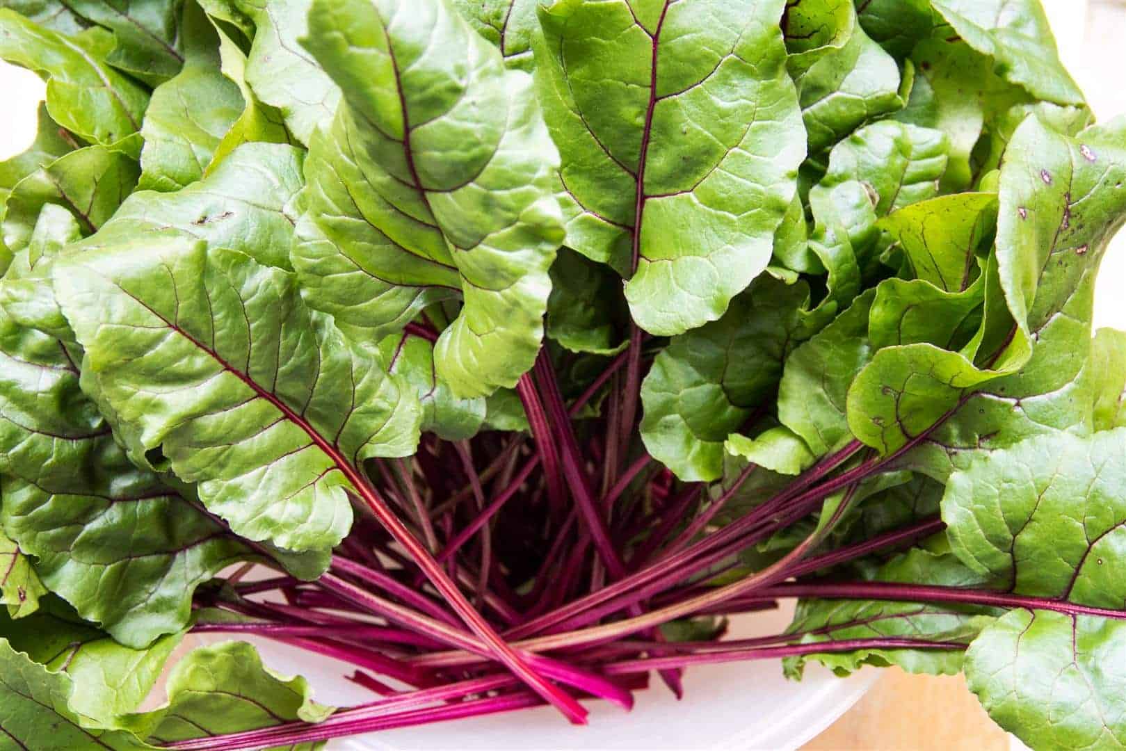 A photo of fresh beetroot leaves, also known as beet greens, arranged in a pile with some of the stems still attached. The leaves have a bright green color and a slightly wrinkled texture, and are presented on a wooden surface