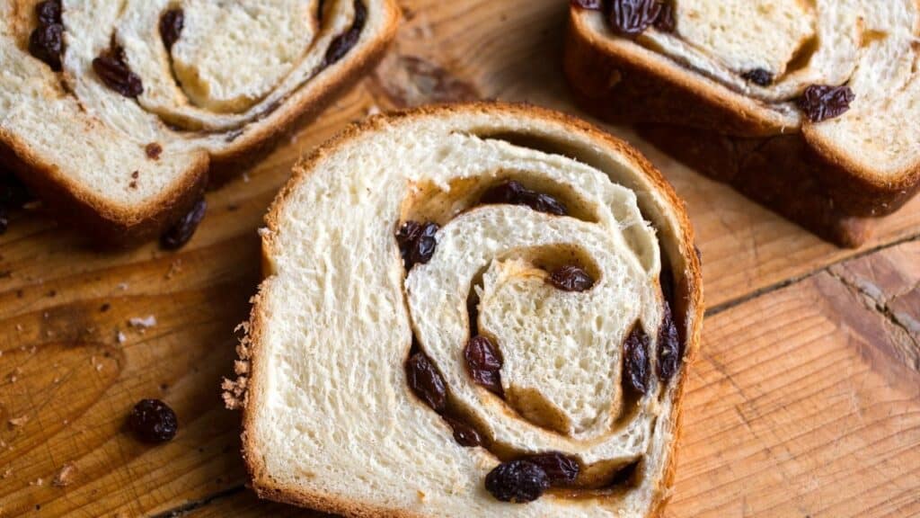 A photo of a freshly baked loaf of cinnamon raisin bread, sliced into thick slices and stacked on a wooden board. 