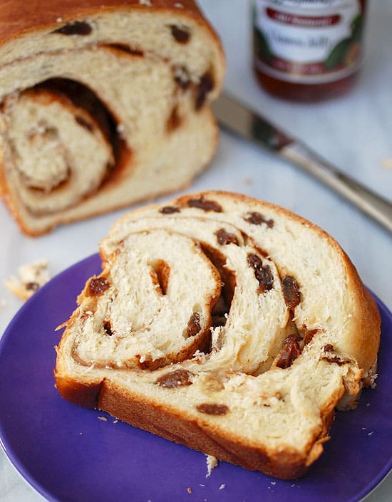 A photo of a freshly baked loaf of cinnamon raisin bread, sliced into thick slices and stacked on a wooden board. The bread has a golden-brown crust and visible raisins and cinnamon swirls throughout.