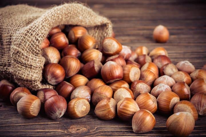 Hazelnuts in shells on wooden surface, oval-shaped with light brown shells, some with green stems.