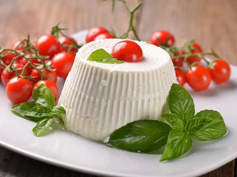 A photo of a dish of creamy ricotta cheese topped with chopped basil, sliced cherry tomatoes, and a drizzle of olive oil.